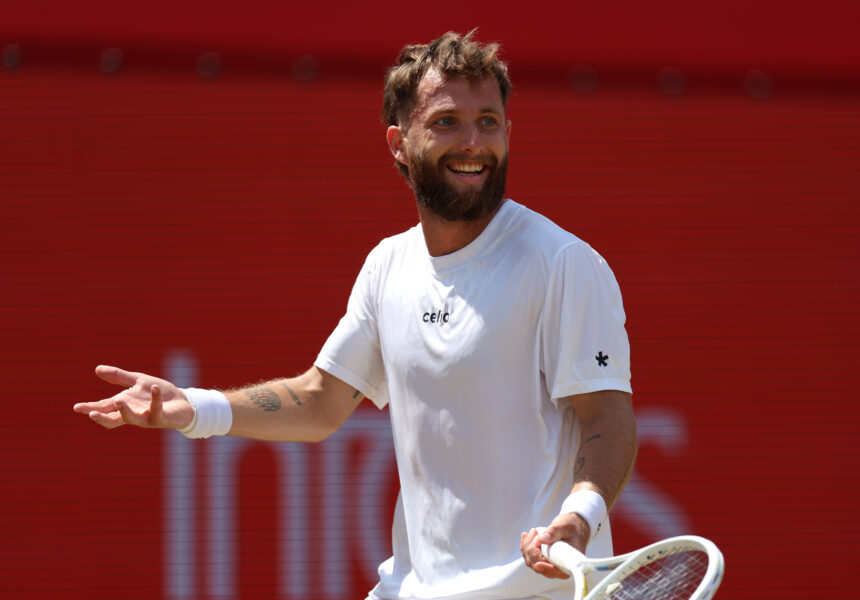 A man in a white shirt smiles while extending his left hand, holding a tennis racket in his right hand against a red background stadium wall.