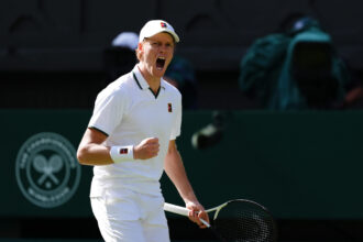 Tennis player in white outfit shouting during a match, holding a racket in front of a dark backdrop, sweat and intensity visible on his face.