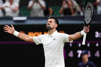 Tennis player in a white shirt raises arms in celebration, holding a racket aloft against a blurred crowd background.