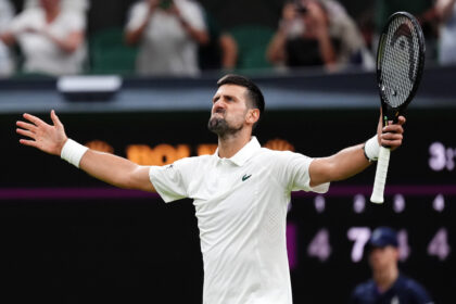 Tennis player in a white shirt raises arms in celebration, holding a racket aloft against a blurred crowd background.