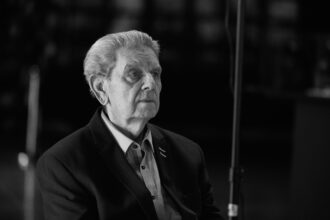 Older man in a blazer and button-down shirt, looking to the left, with a microphone stand in the foreground.