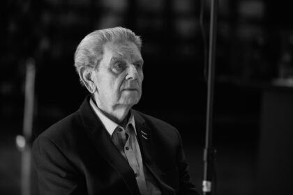 Older man in a blazer and button-down shirt, looking to the left, with a microphone stand in the foreground.