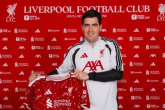 Man in a white Liverpool FC jacket holding a red jersey at a press backdrop featuring LFC logos and sponsors.