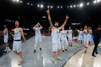 Basketball players in light-blue uniforms celebrate on a court in a packed arena after a game, raising arms in triumph.