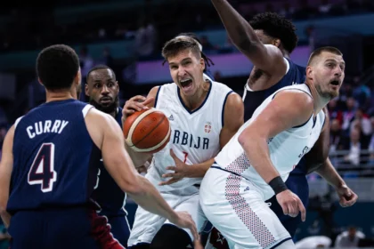 Basketball players in white and blue uniforms battle for the ball in a crowded lane, one player gripping the ball with intense expression as others contest.