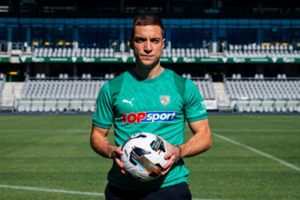 Soccer player in teal jersey holding a white soccer ball on a green field with empty stadium seats behind him.