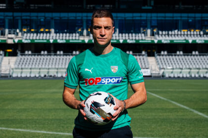 Soccer player in teal jersey holding a white soccer ball on a green field with empty stadium seats behind him.