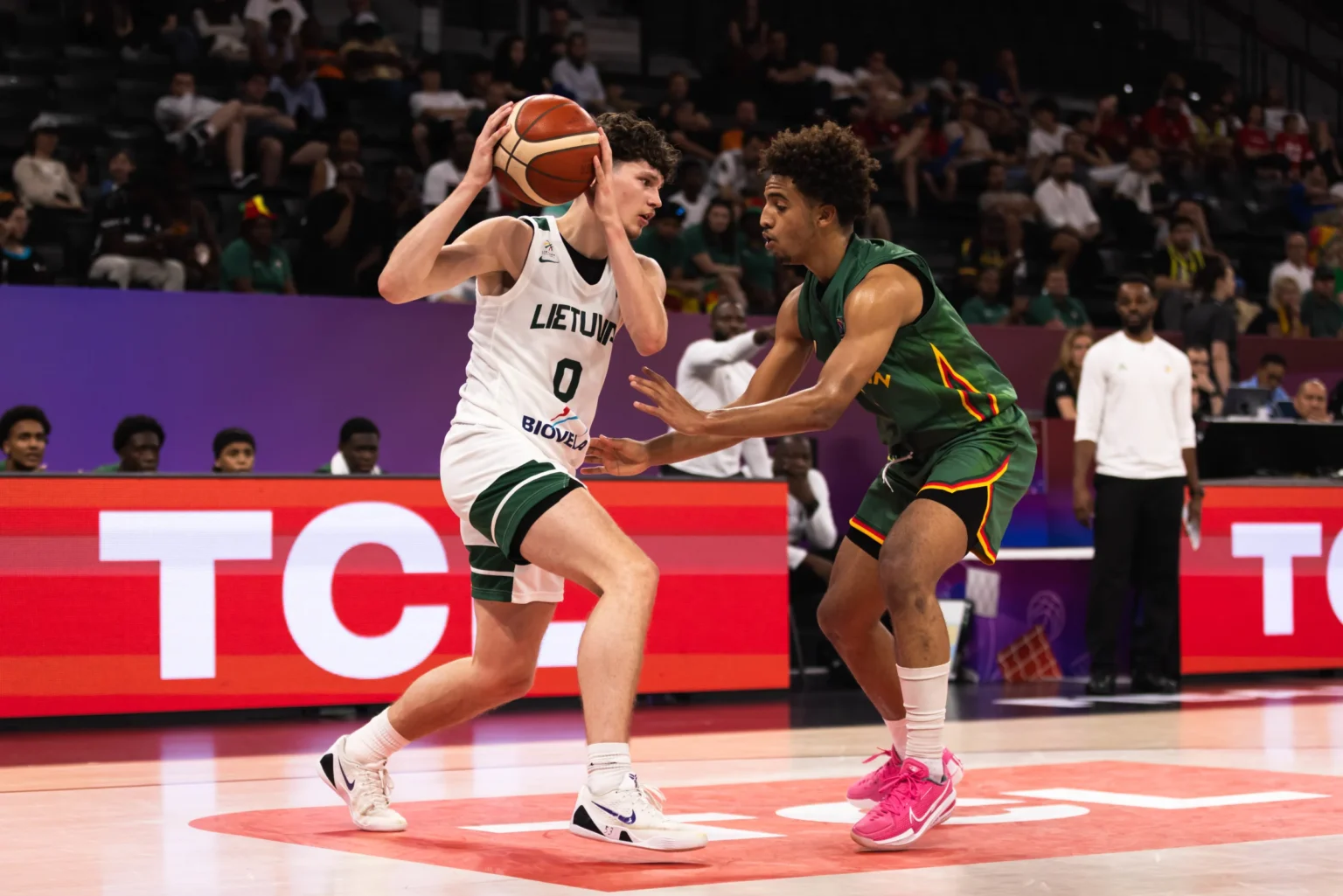 Basketball players contesting a ball near the baseline, Lithuanian white jersey shielding the ball from a defender in green. Crowd in the stands behind.