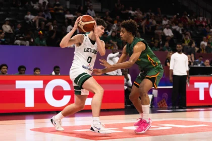 Basketball players contesting a ball near the baseline, Lithuanian white jersey shielding the ball from a defender in green. Crowd in the stands behind.