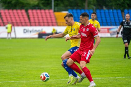 Two soccer players in yellow and red kits chase the ball on a wet, green field with red and blue stadium seats in the background.