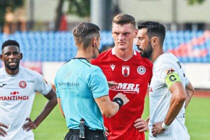 Soccer referee in blue talks to three players on the field during a match, with a central player in red and others in white jerseys nearby.