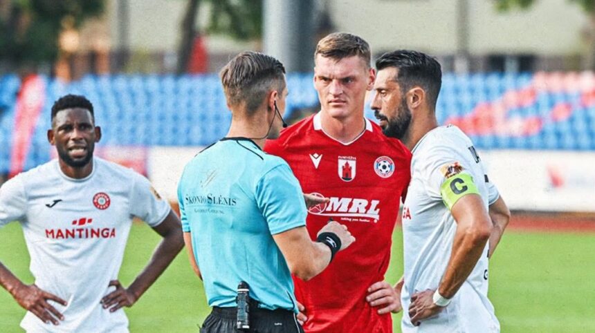 Soccer referee in blue talks to three players on the field during a match, with a central player in red and others in white jerseys nearby.