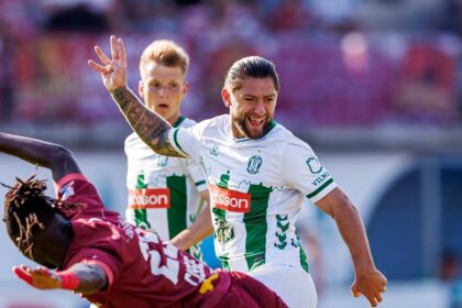 Soccer player in white and green raises his arm in celebration as a teammate approaches on a sunlit field.