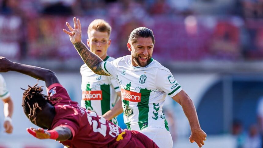 Soccer player in white and green raises his arm in celebration as a teammate approaches on a sunlit field.