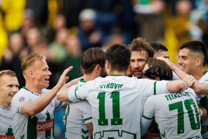 Soccer players in white and green celebrate in a huddle on the field, arms around each other.