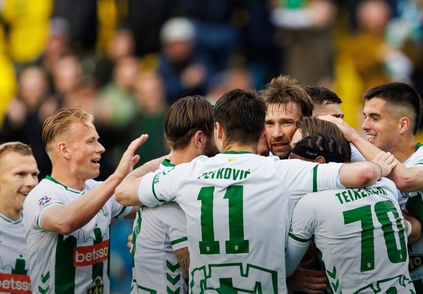 Soccer players in white and green celebrate in a huddle on the field, arms around each other.
