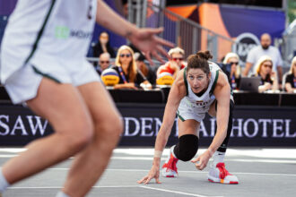 Female volleyball player in white and green crouches for a dig on an indoor court; blurred teammate and spectators in background.