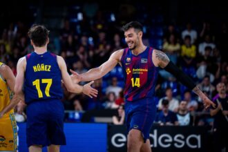 Two basketball players in blue and red jerseys greet each other with a high-five on the court, crowd in the background