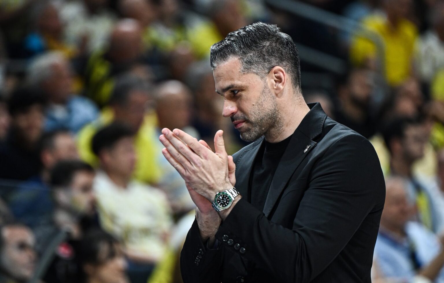 Man in a black suit claps at a stadium, blurred crowd in the background, suggesting applause or celebration.