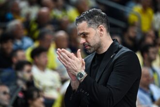 Man in a black suit claps at a stadium, blurred crowd in the background, suggesting applause or celebration.