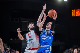 Two basketball players leap for the ball in a crowded arena, contesting a rebound.