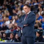 Man in a navy suit with red tie stands with arms crossed at a crowded event or sports arena.