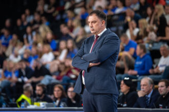 Man in a navy suit with red tie stands with arms crossed at a crowded event or sports arena.