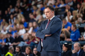 Man in a navy suit with red tie stands with arms crossed at a crowded event or sports arena.