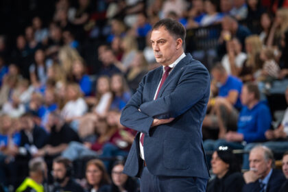 Man in a navy suit with red tie stands with arms crossed at a crowded event or sports arena.
