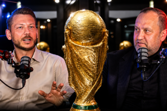 Two men podcasting at a table with a large golden trophy between them on display in a studio setting, speaking into microphones.