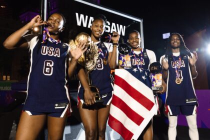 Four USA basketball players pose on a podium at night, wearing navy uniforms with medals, one holding a large torch and another draped in a red-white-and-blue flag.