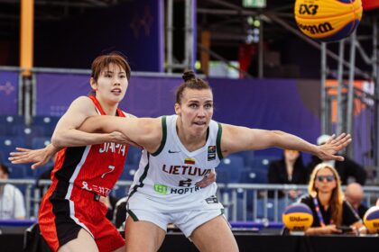 Two female basketball players reach for the ball during a game, one in red and the other in white Lithuania kit with 'LIETUVA' on the jersey.