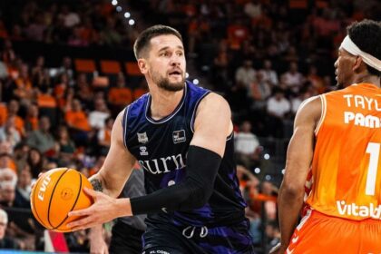 Basketball player in a purple jersey dribbles the ball while a defender in orange guards him on a crowded court.