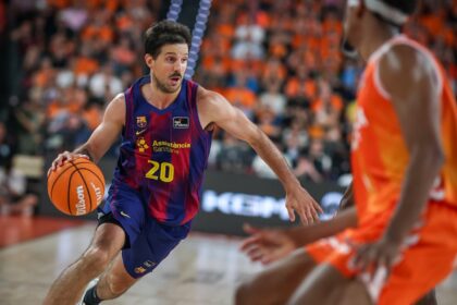 Basketball player in a blue FC Barcelona jersey (#20) dribbles toward the basket as a defender in orange closes in. Crowded arena in background.