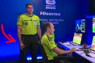 Two referees in neon yellow uniforms stand at a blue VAR review station with multiple screens and game footage on monitors in the background.