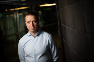 Portrait of a man in a light blue dress shirt leaning against a dark brick wall in a dimly lit corridor, looking toward the camera.