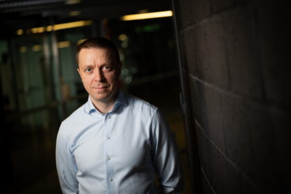 Portrait of a man in a light blue dress shirt leaning against a dark brick wall in a dimly lit corridor, looking toward the camera.