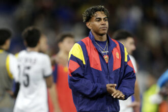 Young man in a red, yellow, and blue track jacket standing on a field with blurred crowd in the background.