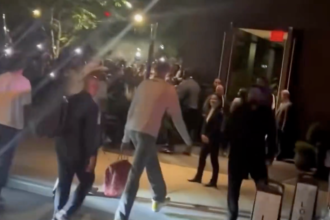 Nighttime street scene showing a crowd gathered outside a doorway, with a man in light-colored jeans walking toward the entrance and colorful lights in the background.