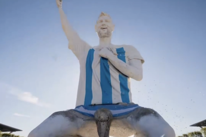 Giant statue of a man in a blue-and-white striped robe, right arm raised, outdoors with a cheering crowd below.