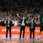 Coaches and players in suits raise arms on a basketball court as a cheering crowd in green fills the stands behind them.