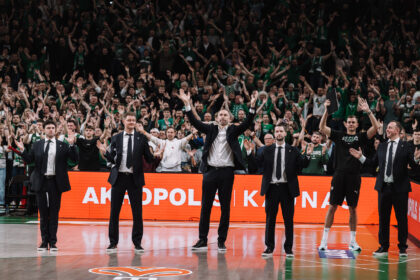 Coaches and players in suits raise arms on a basketball court as a cheering crowd in green fills the stands behind them.