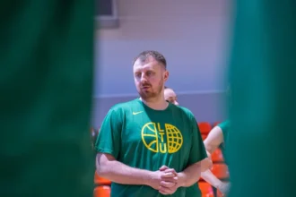 Man wearing a green T-shirt with a yellow basketball-like logo stands in an indoor gym, hands clasped, orange seats in the background