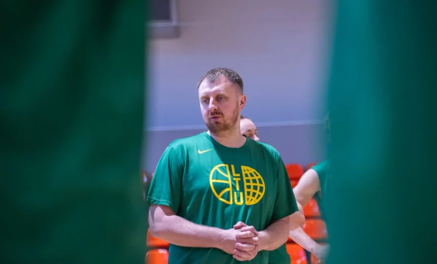 Man wearing a green T-shirt with a yellow basketball-like logo stands in an indoor gym, hands clasped, orange seats in the background
