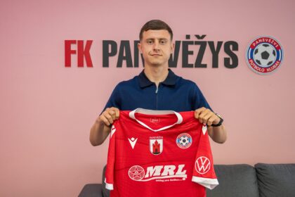 Man in a blue polo presenting a red soccer jersey with Panevėžys badges, against a pink wall with a circular club logo to the right.