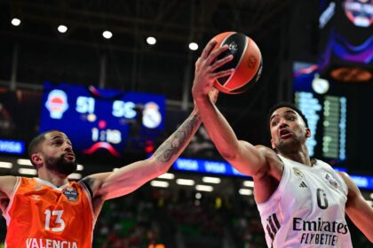 Two basketball players reach for the ball mid-air: Valencia in orange 13 on the left and Real Madrid in white on the right, with arena lights in the background to indicate a game.