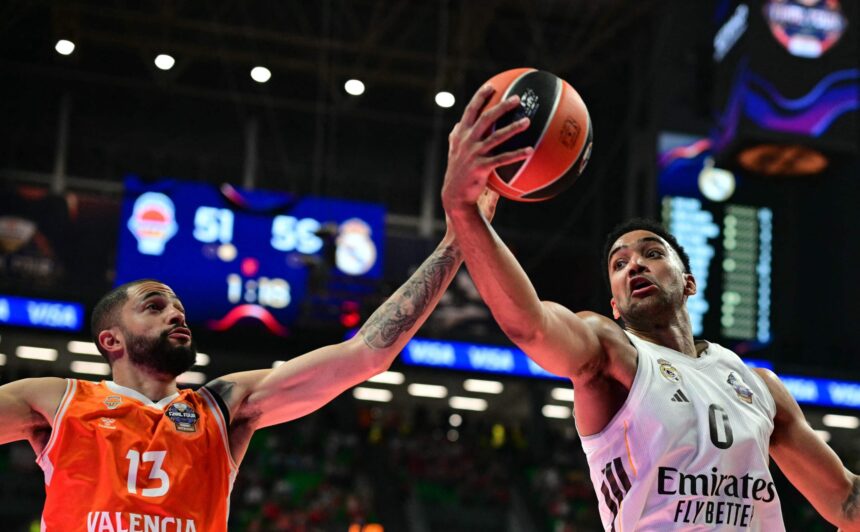 Two basketball players reach for the ball mid-air: Valencia in orange 13 on the left and Real Madrid in white on the right, with arena lights in the background to indicate a game.