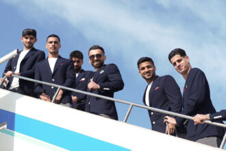 Six men in navy blazers posing on a yacht deck against a blue sky.