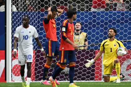 Soccer scene: goalkeeper in yellow kneels with ball near the goal as players in red and blue jerseys stand nearby behind the net.