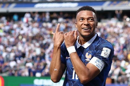 Soccer player in blue France jersey celebrating on the field with hands raised, crowd in the stands behind him.